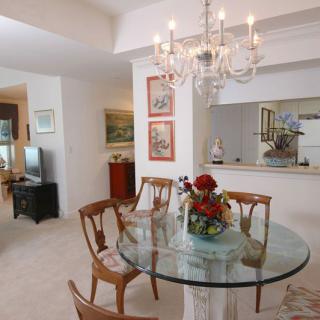 A cozy dining room with a glass table and wooden chairs, illuminated by a chandelier and decorated with flowers.