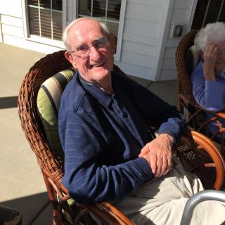 An elderly man is smiling while sitting outdoors on a sunny patio in a wicker chair.