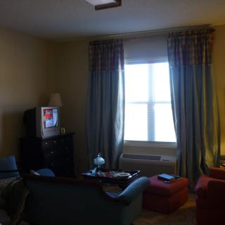 A cozy living room with a television, sofa, and chairs near a curtained window.