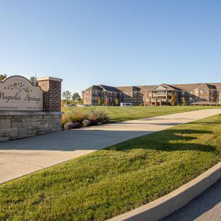 A view of a residential apartment complex with a large green lawn and a welcoming driveway entrance.