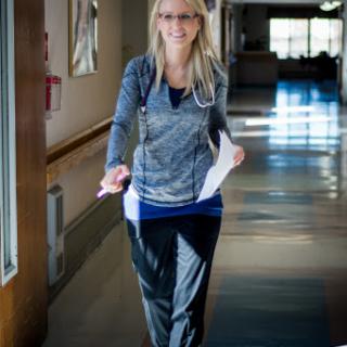 A nurse in scrubs walks through a hospital corridor, holding paperwork and wearing a stethoscope.