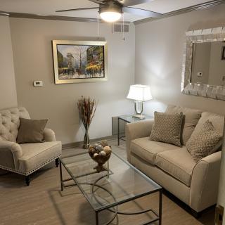 A cozy living room with elegant furniture and decor featuring a sofa, armchair, glass coffee table, lamp, wall art, and a mirror.