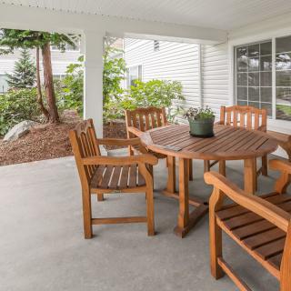 A cozy outdoor patio area with a round wooden table and matching chairs under a covered porch, overlooking a garden.