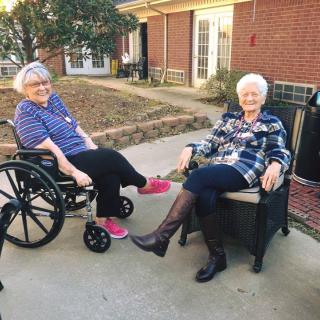 Two elderly women enjoying a sunny day outdoors, one in a wheelchair and the other seated in a chair by a brick building.