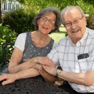 An elderly couple sitting together outdoors, smiling warmly at the camera.