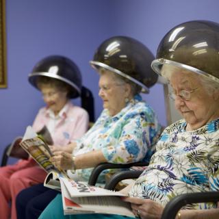 Elderly women sitting under hair dryers and reading in a beauty salon.