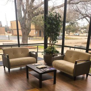 A cozy lobby seating area with two armchairs, a table, and a plant by large windows overlooking the outside.