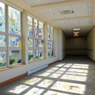 A brightly lit hallway featuring large stained glass windows that create intricate patterns of light and shadow on the floor.