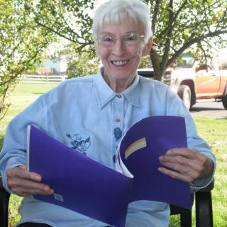 An elderly person sitting outdoors, enjoying reading a book and smiling.