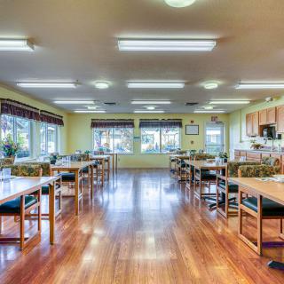 A spacious dining area with wooden flooring and ample natural light, featuring multiple tables and a kitchen area.
