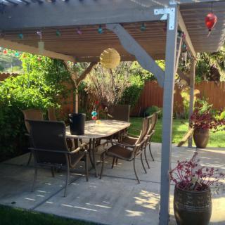 A sunny patio with a pergola, table, and chairs surrounded by a lush garden.