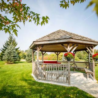 A serene gazebo in a lush green park, surrounded by trees and flowers under a clear blue sky.