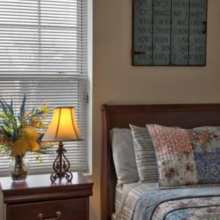 A cozy bedroom corner with a neatly made bed, nightstand, lamp, and decorative flowers by the window.
