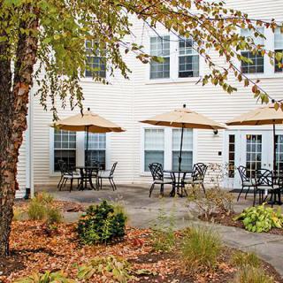An outdoor patio area with tables and umbrellas in front of a building surrounded by landscaped greenery.