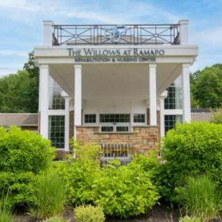 A front view of a building entrance with a sign reading 'The Willows at Elmwood' surrounded by landscaped gardens.