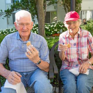 Two elderly people enjoying a snack while sitting on a park bench.