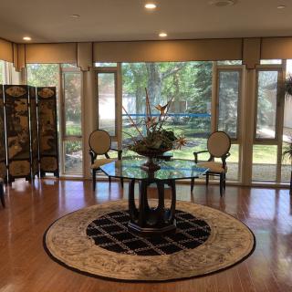 A stylish living room with a glass table centerpiece, surrounded by chairs and large windows allowing ample natural light.
