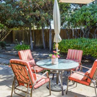 A cozy patio setting with a round table, umbrella, and four cushioned chairs in a lush backyard.