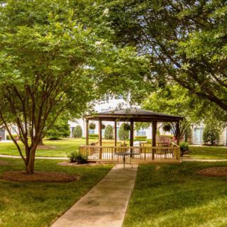 A peaceful park setting featuring a wooden gazebo surrounded by lush trees and greenery along a concrete pathway.