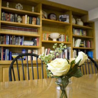 A cozy dining room setting featuring a vase of white roses on a polished table, with shelves filled with books and decorative items in the background.