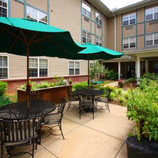 A cozy outdoor patio area with tables, chairs, and green umbrellas surrounded by lush greenery in a modern residential complex.