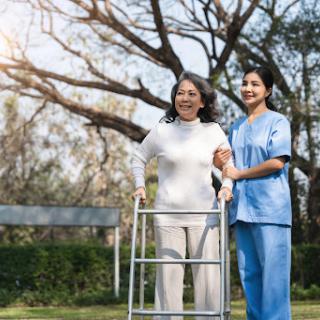 A nurse assists an elderly woman using a walker in a serene outdoor setting.