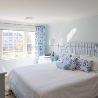 A bright and cozy bedroom with a light blue color scheme, featuring a bed adorned with decorative pillows and plenty of natural sunlight.