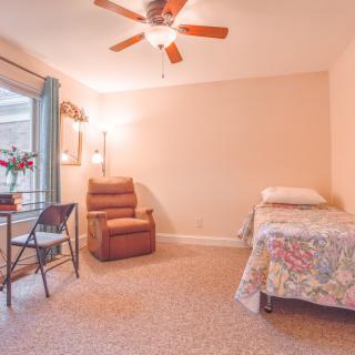 A cozy bedroom with a single bed, armchair, and folding table, featuring a ceiling fan and warm lighting.