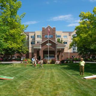 A group of people enjoying a sunny day on the lawn in front of a multi-story building surrounded by trees.