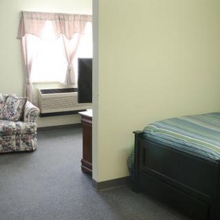 A simple hotel room with a bed and living area, featuring a TV and an armchair near the window.