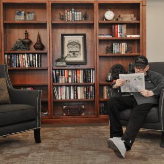 A person sitting in an armchair reads a newspaper in front of a well-stocked bookshelf.