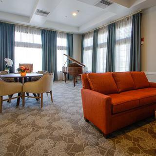 A spacious living room featuring an orange sofa, a grand piano, and a dining table with chairs, set against large windows with flowing curtains and a patterned carpet.