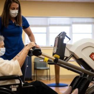 A healthcare professional assists an elderly person in a wheelchair with exercise equipment in a rehabilitation setting, both wearing masks.
