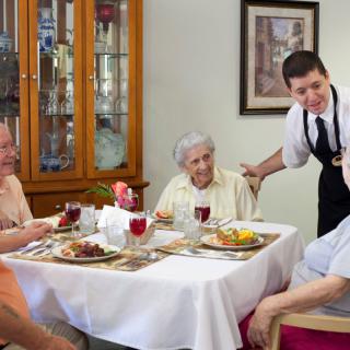 Seniors enjoying a meal together in a dining setting with a waiter attending to them.
