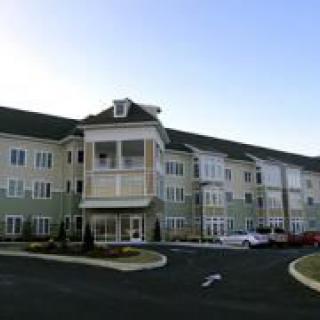 A large residential apartment complex with multiple windows, featuring three stories and a parking area in front.