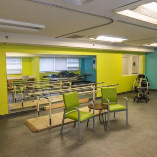 A brightly colored rehabilitation room equipped with parallel bars, a mirror, and chairs for physical therapy.