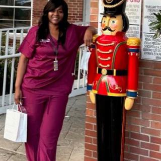 A person in scrubs poses next to a large decorative nutcracker statue outdoors.