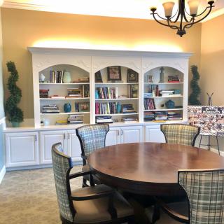 A cozy dining room with a round table, surrounded by chairs and a set of bookshelves in the background.