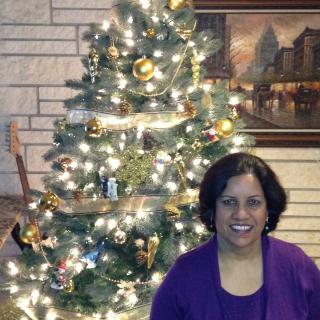 A woman smiling in front of a decorated Christmas tree adorned with lights and ornaments inside a cozy home.