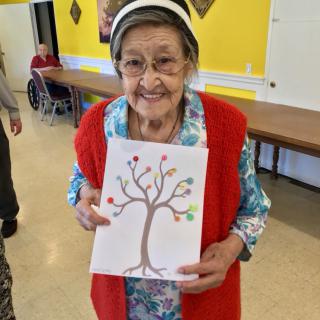 An elderly woman smiling and holding a drawing of a tree indoors.