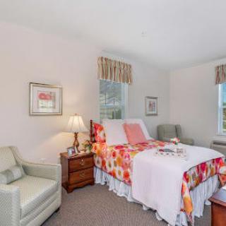 A cozy bedroom interior with a bed adorned in floral bedding, a lamp on a nightstand, and comfortable seating by the window.