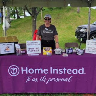 A volunteer stands at a Home Instead booth during a community event, providing information and resources.