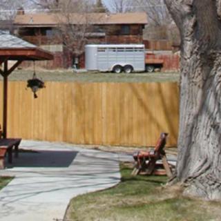 A backyard scene with a wooden fence, large tree, and a bench on a paved path. A trailer is visible in the background.
