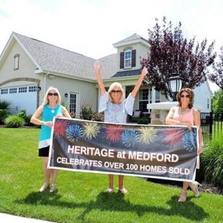 Three people celebrating in front of a house with a banner reading 'Heritage at Medford Celebrates Over 100 Homes Sold.'