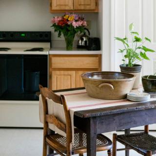 A cozy kitchen with a wooden table and chairs, a large bowl, indoor plants, and flowers near the stove.