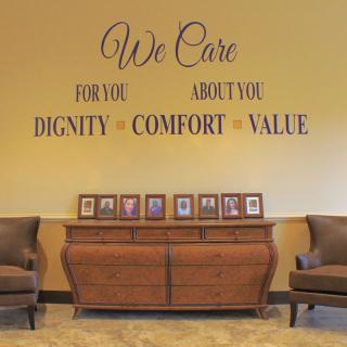 A warmly decorated room featuring a wooden dresser with framed photos, flanked by two chairs. The wall displays inspirational phrases: 'We Care For You About You - Dignity Comfort Value.'