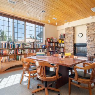 A cozy library room with large windows, a wooden table surrounded by chairs, and bookshelves lining the walls.