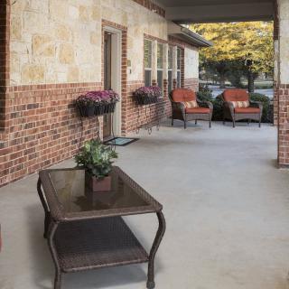 A cozy porch with potted plants, chairs, and a table against a brick wall.