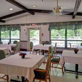 A well-lit dining room featuring tables with white tablecloths and chairs, framed by large windows and decorative elements.