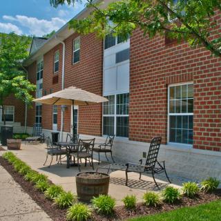 A brick building with a patio area featuring outdoor seating and an umbrella, surrounded by landscaping.